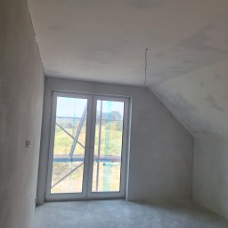 USŁUGI OGÓLNOBUDOWLANE MARCIN MISZK - Poddasze w stanie deweloperskim z oknem balkonowym, ściany i sufit pokryte szarą warstwą gładzi, widoczne kable wystające z sufitu, przez okno widoczny krajobraz i elementy rusztowania.