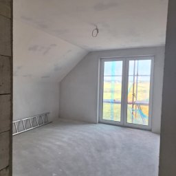 USŁUGI OGÓLNOBUDOWLANE MARCIN MISZK - Poddasze w stanie surowym, z widocznymi śladami po gładzi na ścianach i suficie, aluminiowa drabina oparta o ścianę, okno zabezpieczone folią malarską, surowy beton na podłodze.