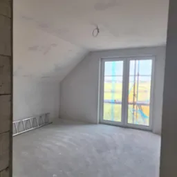 Poddasze w stanie surowym, z widocznymi śladami po gładzi na ścianach i suficie, aluminiowa drabina oparta o ścianę, okno zabezpieczone folią malarską, surowy beton na podłodze.