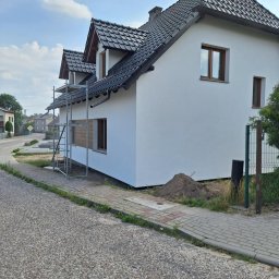 USŁUGI OGÓLNOBUDOWLANE MARCIN MISZK - Dom jednorodzinny z nową, białą elewacją, widoczne rusztowanie przy bocznej ścianie, okna zabezpieczone folią, niewielka górka ziemi przy ogrodzeniu.