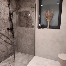 USŁUGI OGÓLNOBUDOWLANE MARCIN MISZK - Nowoczesna kabina prysznicowa z deszczownicą w łazience wykończonej marmurowymi płytkami, dekorowana suszonymi trawami w wazonie na parapecie okna.