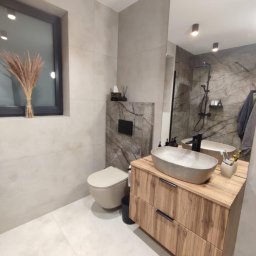 USŁUGI OGÓLNOBUDOWLANE MARCIN MISZK - Nowoczesna łazienka z beżowymi płytkami, wiszącą toaletą, drewnianą szafką pod umywalkę z szarą, owalną umywalką nablatową i dużym lustrem odbijającym prysznic.