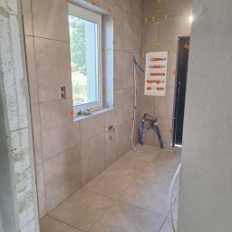 USŁUGI OGÓLNOBUDOWLANE MARCIN MISZK - Beżowe płytki na ścianach i podłodze w trakcie montażu, widoczne krzyżyki dystansowe, okno z widokiem na zieleń, przygotowana instalacja wodna i tablica rozdzielcza.