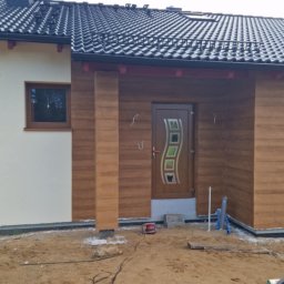 USŁUGI OGÓLNOBUDOWLANE MARCIN MISZK - Fragment elewacji domu jednorodzinnego z wykończonym wejściem, drewnopodobna okładzina, widoczne okna z brązowymi ramami, dachówka ceramiczna, teren wokół w trakcie prac wykończeniowych.