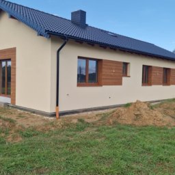 USŁUGI OGÓLNOBUDOWLANE MARCIN MISZK - Wykończony dom jednorodzinny z beżową elewacją, drewnianymi obramowaniami okien i ciemnym dachem, widoczny z perspektywy ogrodu z pryzmami ziemi.