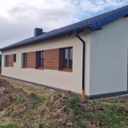 USŁUGI OGÓLNOBUDOWLANE MARCIN MISZK - Widok z zewnątrz na dom jednorodzinny z beżową elewacją i ciemnymi drewnianymi ramami okiennymi, z widoczną stertą ziemi wzdłuż fundamentów.