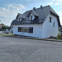 USŁUGI OGÓLNOBUDOWLANE MARCIN MISZK - Dom jednorodzinny z nową, białą elewacją i ciemną dachówką, na tle błękitnego nieba z chmurami; widoczne rusztowania przy oknach na parterze, sugerujące trwające prace wykończeniowe.