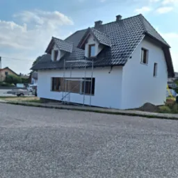 Dom jednorodzinny z nową, białą elewacją i ciemną dachówką, na tle błękitnego nieba z chmurami; widoczne rusztowania przy oknach na parterze, sugerujące trwające prace wykończeniowe.