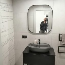 USŁUGI OGÓLNOBUDOWLANE MARCIN MISZK - Nowoczesna łazienka z szarą umywalką nablatową i czarną armaturą, widoczna w lustrze osoba robiąca zdjęcie, ściany wyłożone płytkami imitującymi marmur.