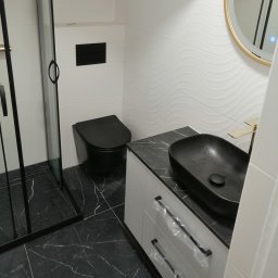 USŁUGI OGÓLNOBUDOWLANE MARCIN MISZK - Nowoczesna łazienka z czarną ceramiką: toaleta, umywalka na blacie imitującym marmur, złota bateria i kabina prysznicowa z czarnymi ramami.