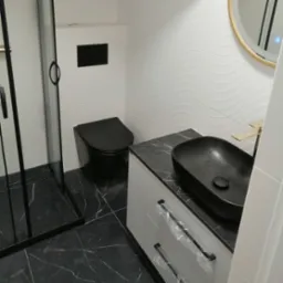 Nowoczesna łazienka z czarną ceramiką: toaleta, umywalka na blacie imitującym marmur, złota bateria i kabina prysznicowa z czarnymi ramami.