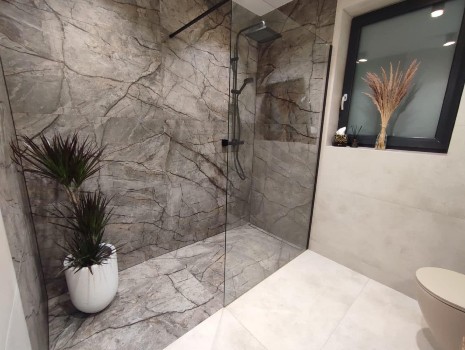 Nowoczesna kabina prysznicowa walk-in z szarymi płytkami imitującymi marmur, czarną armaturą i rośliną doniczkową w białej osłonce, uzupełniona dekoracją w oknie.