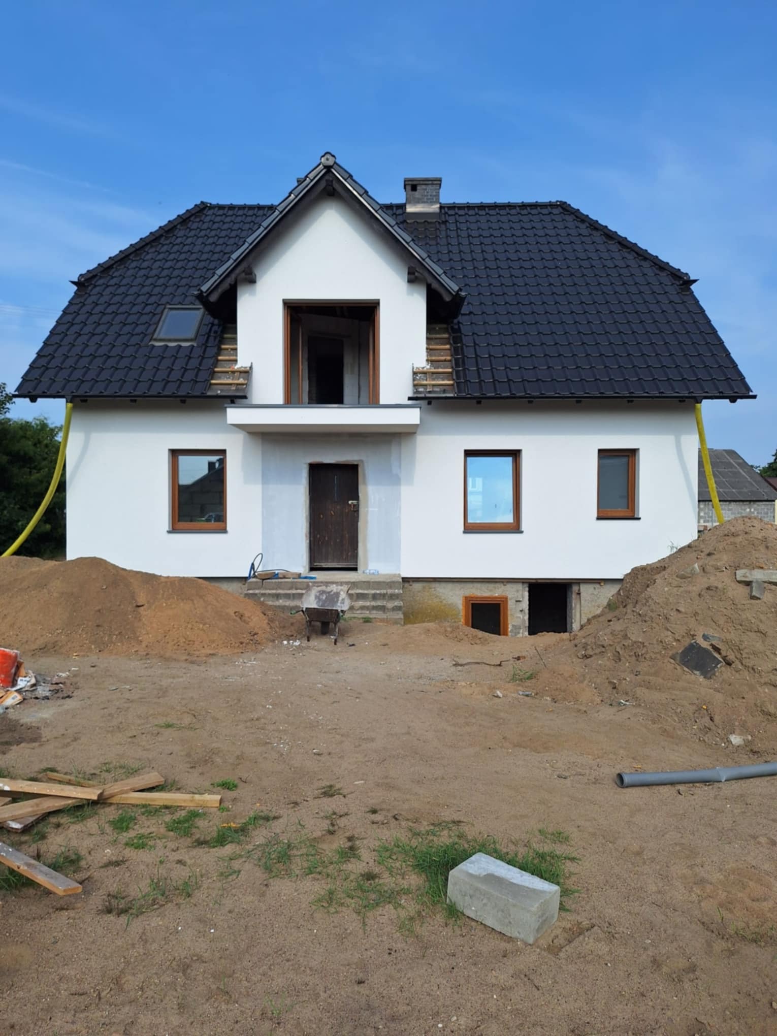 Dom jednorodzinny w trakcie ocieplania elewacji, widoczne rusztowania pod dachem, sterta piasku przed budynkiem i żółty wąż doprowadzający materiał.