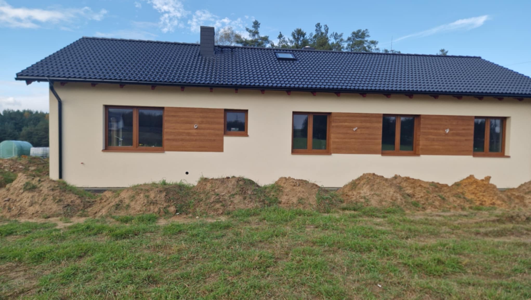 Nowo wybudowany dom jednorodzinny z beżową elewacją, ciemną dachówką i brązowymi oknami, widoczny z poziomu trawnika z usypanymi kopcami ziemi.