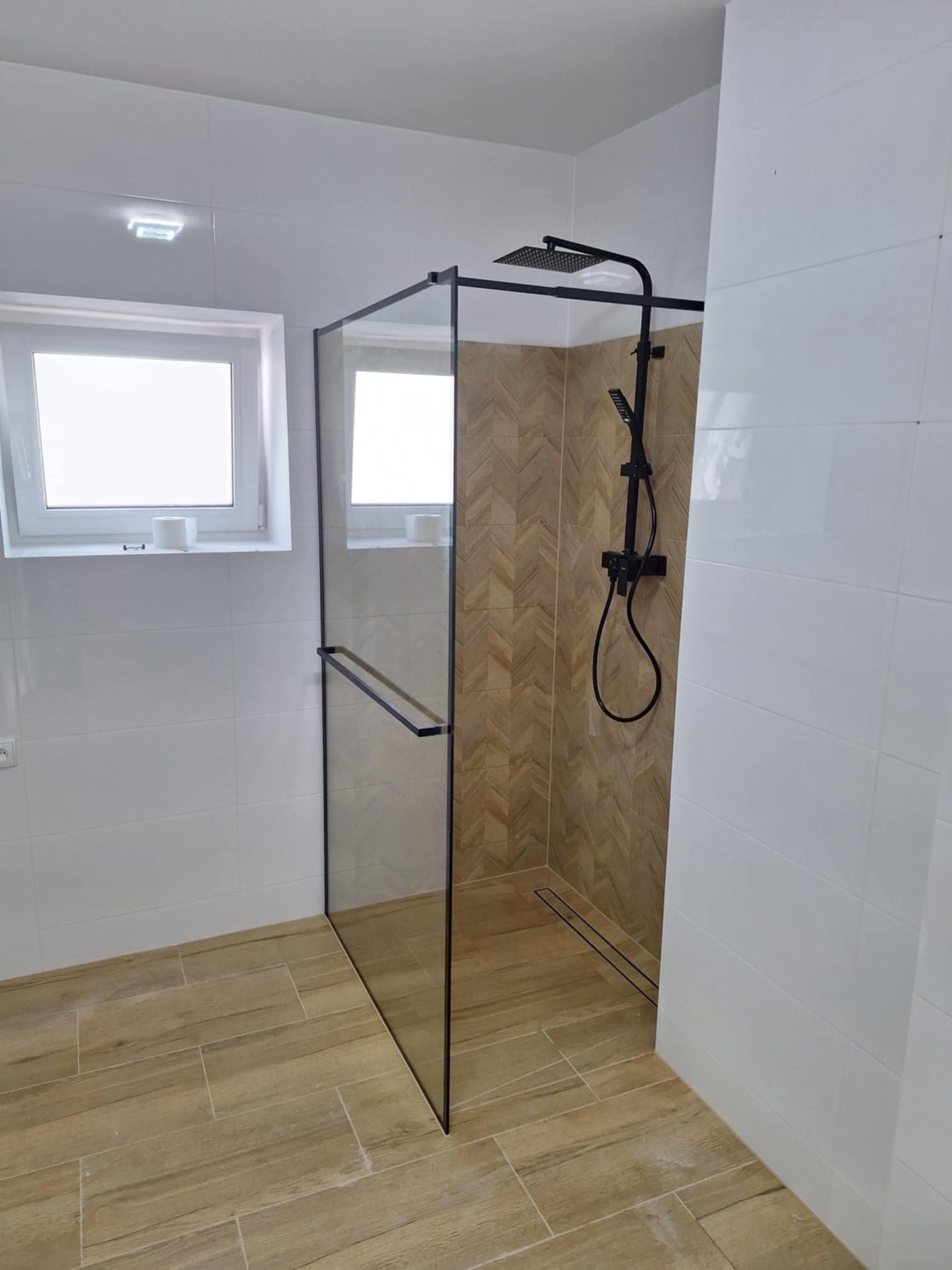 Nowoczesna kabina prysznicowa typu walk-in z czarną armaturą i odpływem liniowym, wykończona białymi płytkami i drewnopodobnymi płytkami ułożonymi w jodełkę wewnątrz strefy prysznicowej.