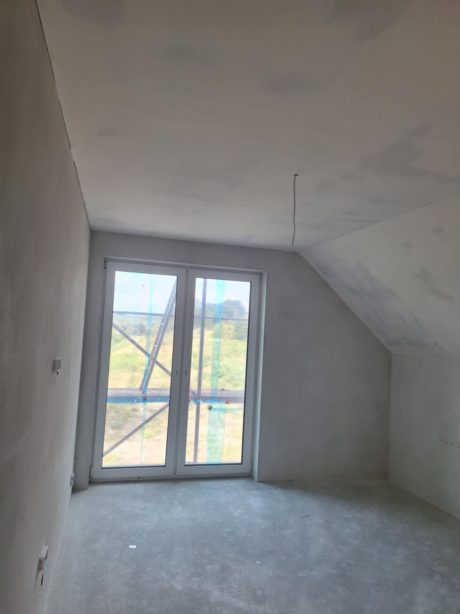 Poddasze w stanie deweloperskim z oknem balkonowym, ściany i sufit pokryte szarą warstwą gładzi, widoczne kable wystające z sufitu, przez okno widoczny krajobraz i elementy rusztowania.