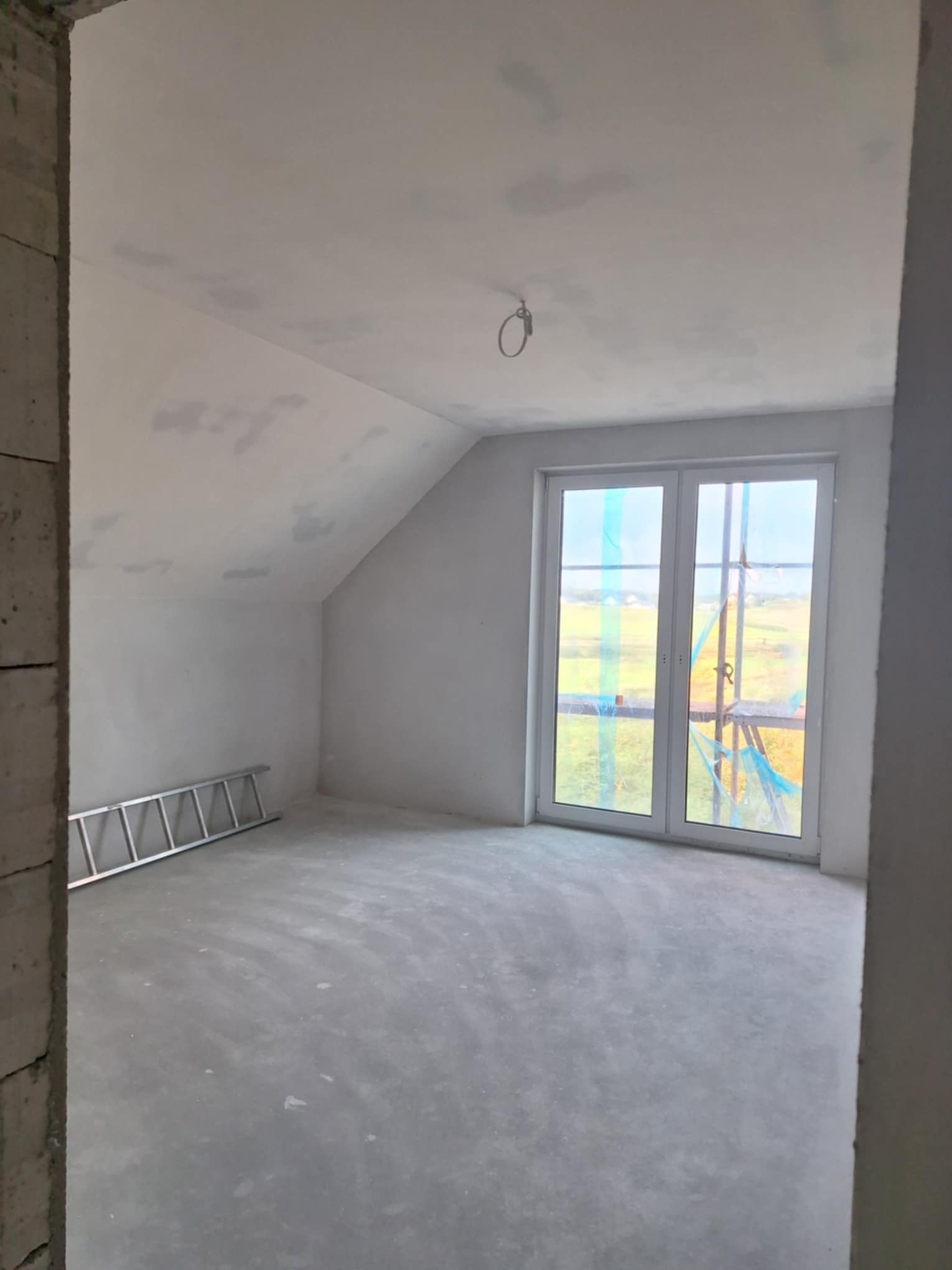 Poddasze w stanie surowym, z widocznymi śladami po gładzi na ścianach i suficie, aluminiowa drabina oparta o ścianę, okno zabezpieczone folią malarską, surowy beton na podłodze.