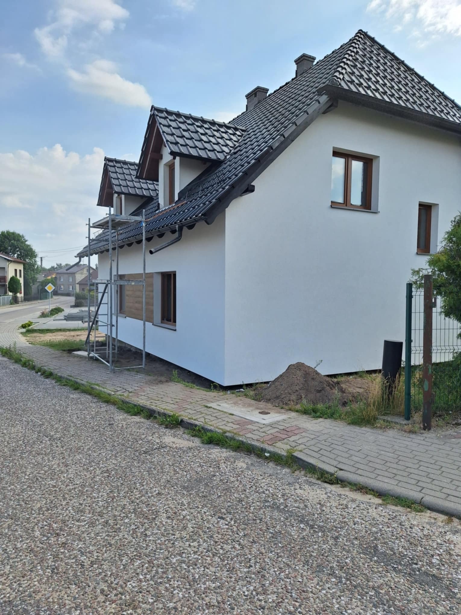 Dom jednorodzinny z nową, białą elewacją, widoczne rusztowanie przy bocznej ścianie, okna zabezpieczone folią, niewielka górka ziemi przy ogrodzeniu.