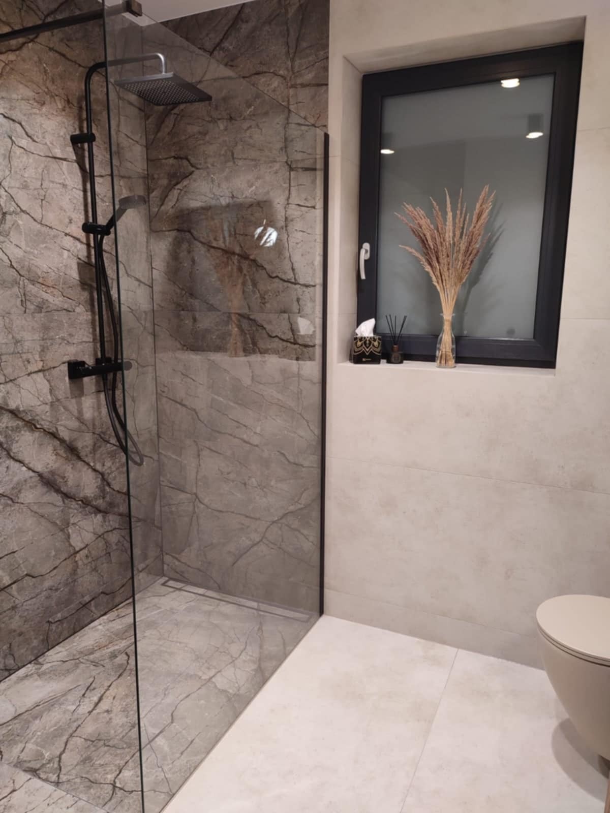 Nowoczesna kabina prysznicowa z deszczownicą w łazience wykończonej marmurowymi płytkami, dekorowana suszonymi trawami w wazonie na parapecie okna.