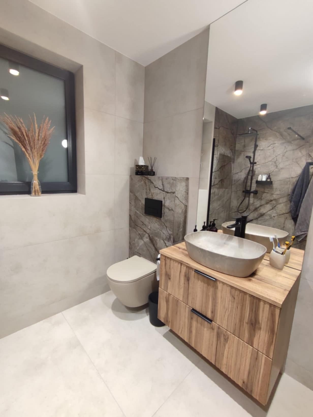 Nowoczesna łazienka z beżowymi płytkami, wiszącą toaletą, drewnianą szafką pod umywalkę z szarą, owalną umywalką nablatową i dużym lustrem odbijającym prysznic.