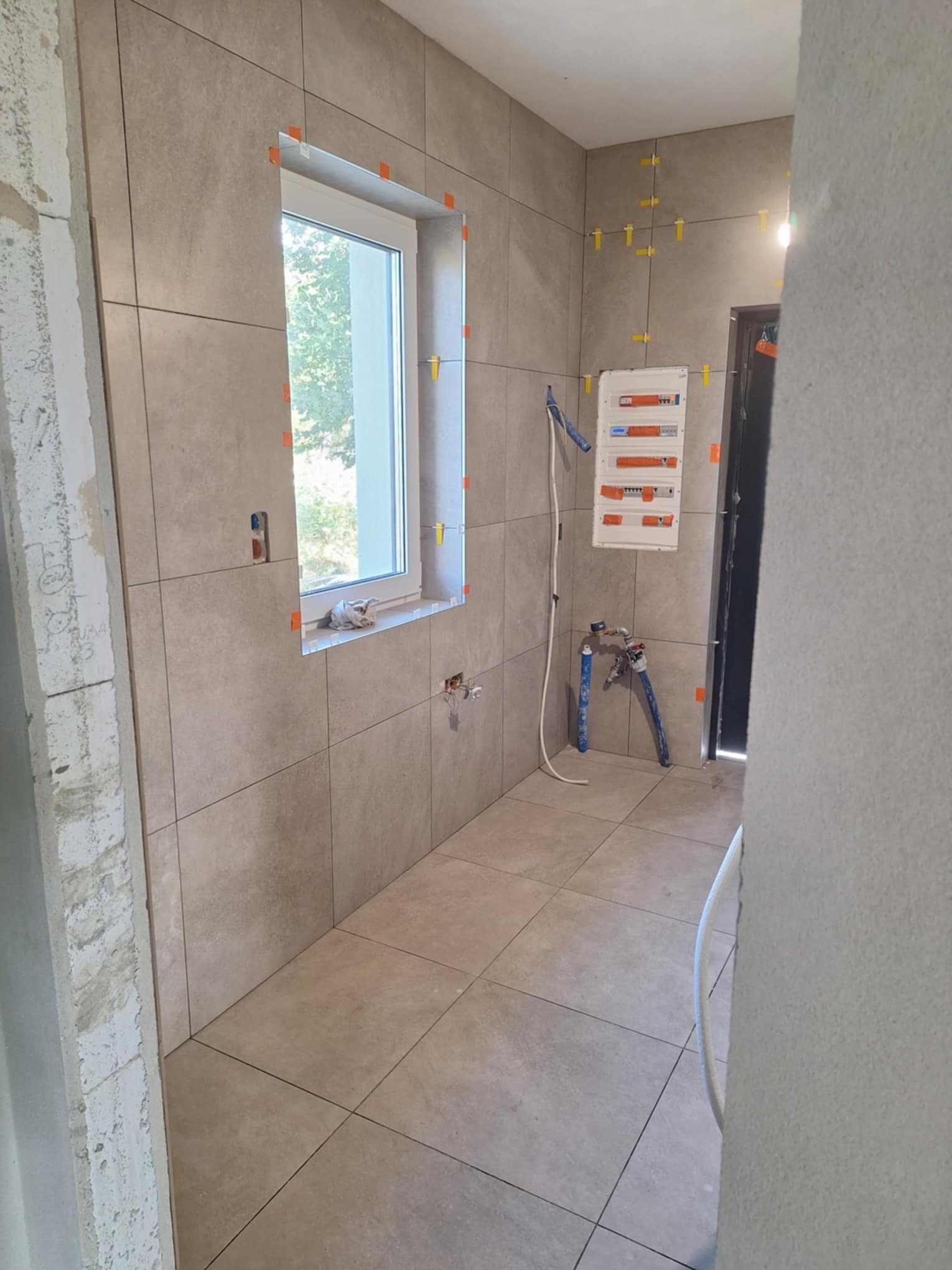 Beżowe płytki na ścianach i podłodze w trakcie montażu, widoczne krzyżyki dystansowe, okno z widokiem na zieleń, przygotowana instalacja wodna i tablica rozdzielcza.
