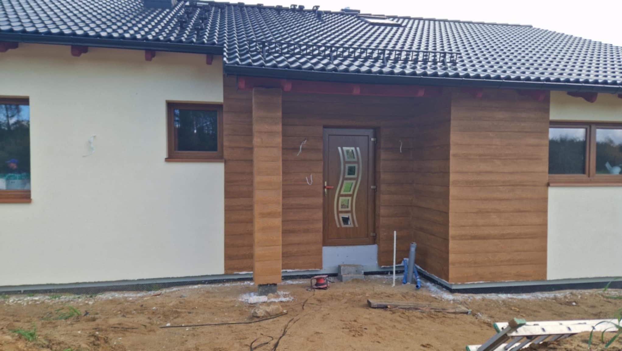 Fragment elewacji domu jednorodzinnego z wykończonym wejściem, drewnopodobna okładzina, widoczne okna z brązowymi ramami, dachówka ceramiczna, teren wokół w trakcie prac wykończeniowych.