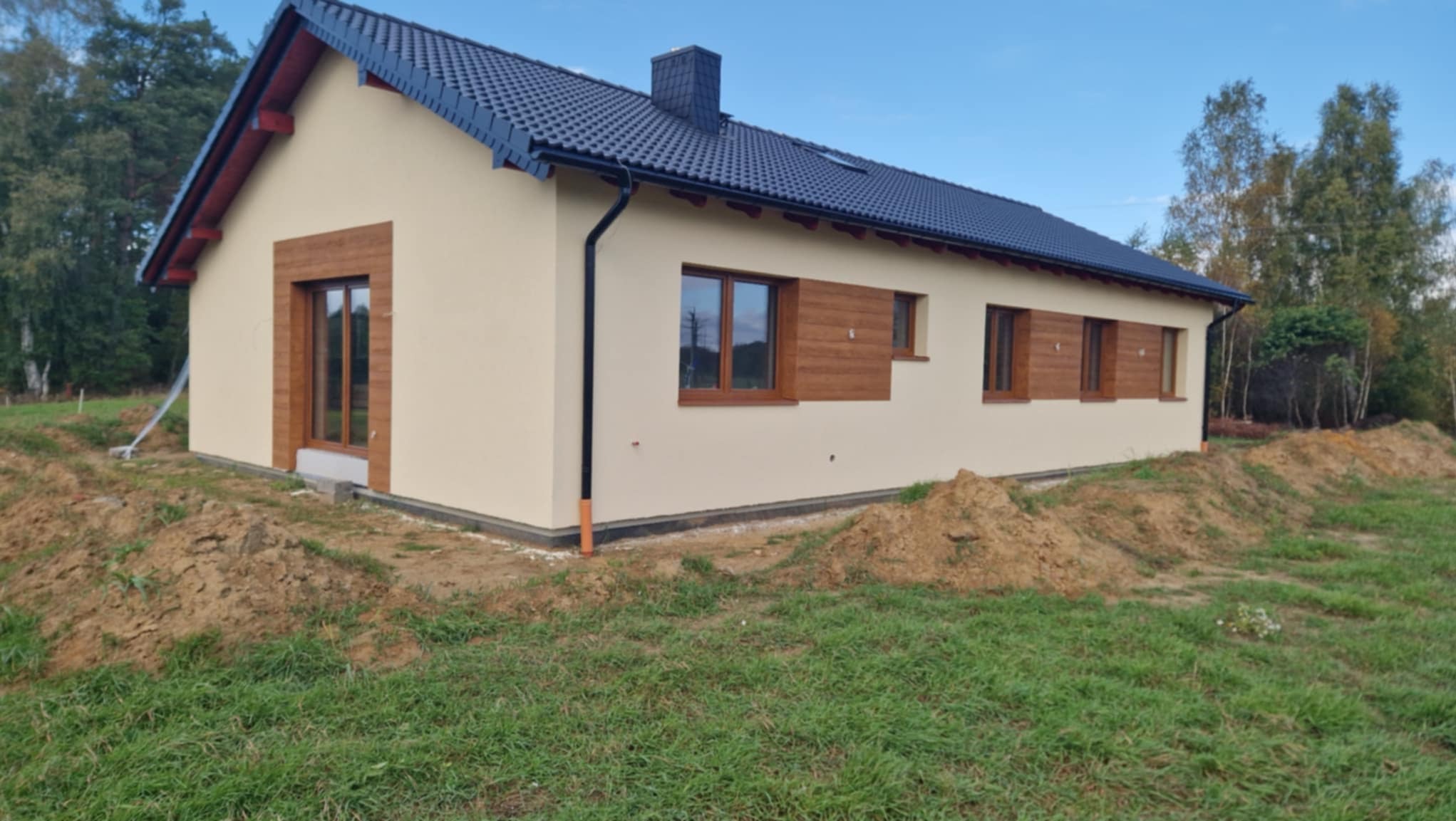 Wykończony dom jednorodzinny z beżową elewacją, drewnianymi obramowaniami okien i ciemnym dachem, widoczny z perspektywy ogrodu z pryzmami ziemi.