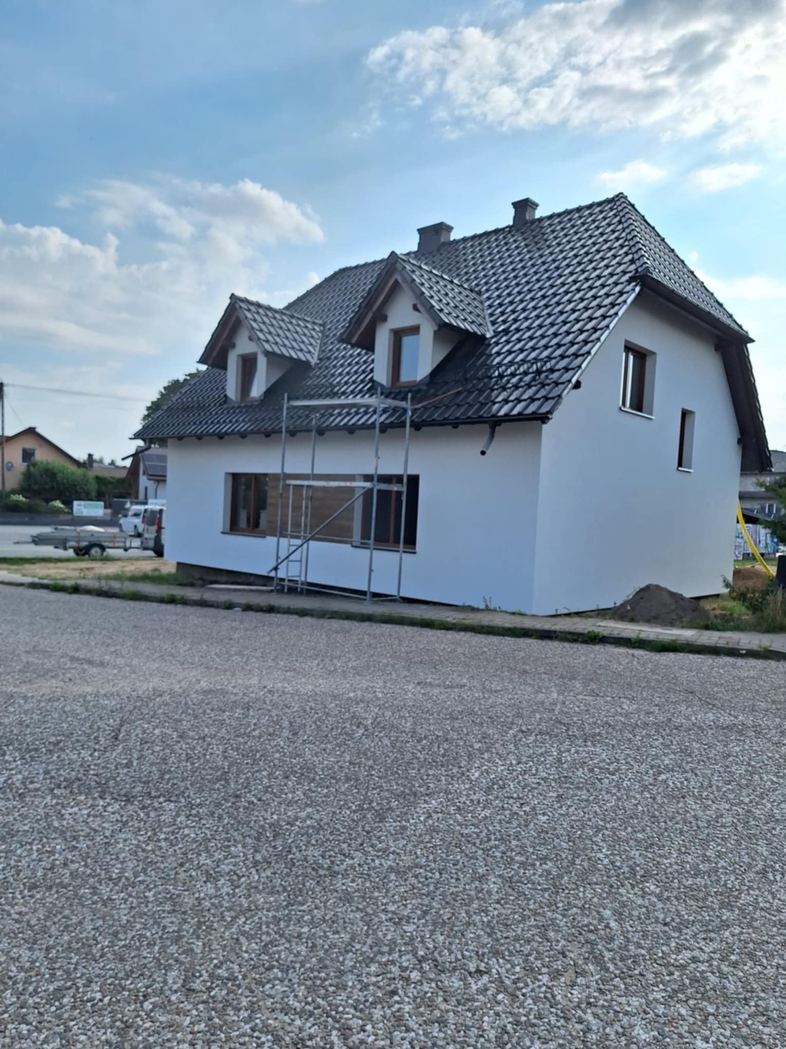 Dom jednorodzinny z nową, białą elewacją i ciemną dachówką, na tle błękitnego nieba z chmurami; widoczne rusztowania przy oknach na parterze, sugerujące trwające prace wykończeniowe.