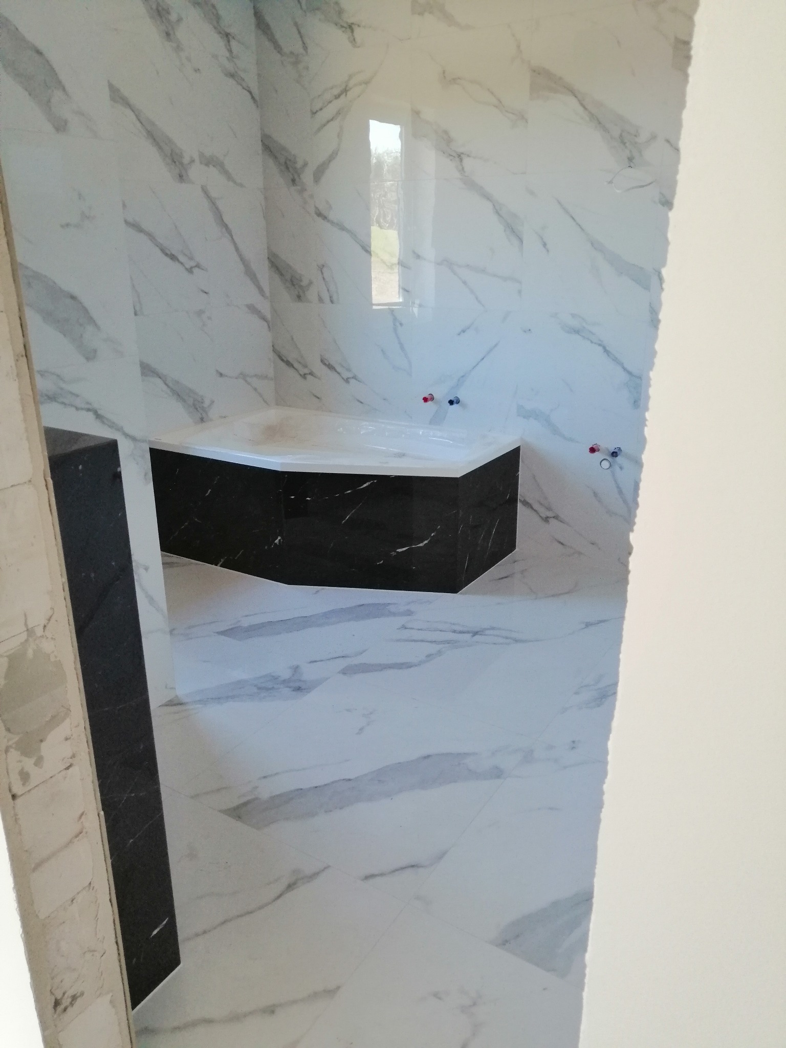 Narożna wanna z czarną obudową w łazience wyłożonej białymi płytkami imitującymi marmur, widoczne wystające rury instalacji wodnej.