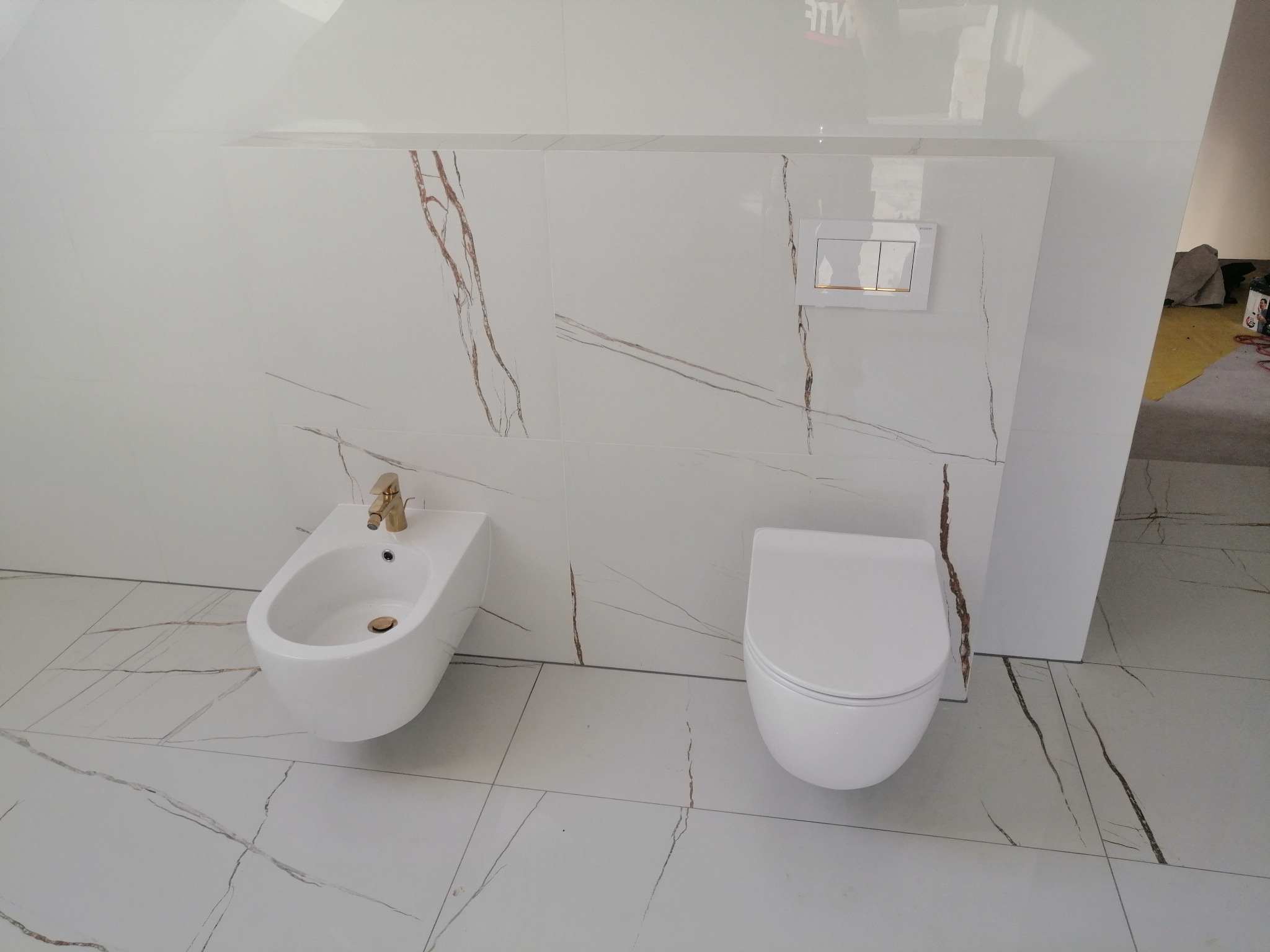 Biała łazienka z płytkami imitującymi marmur, wiszący bidet z złotą baterią i toaleta podwieszana z chromowanym przyciskiem spłukiwania.