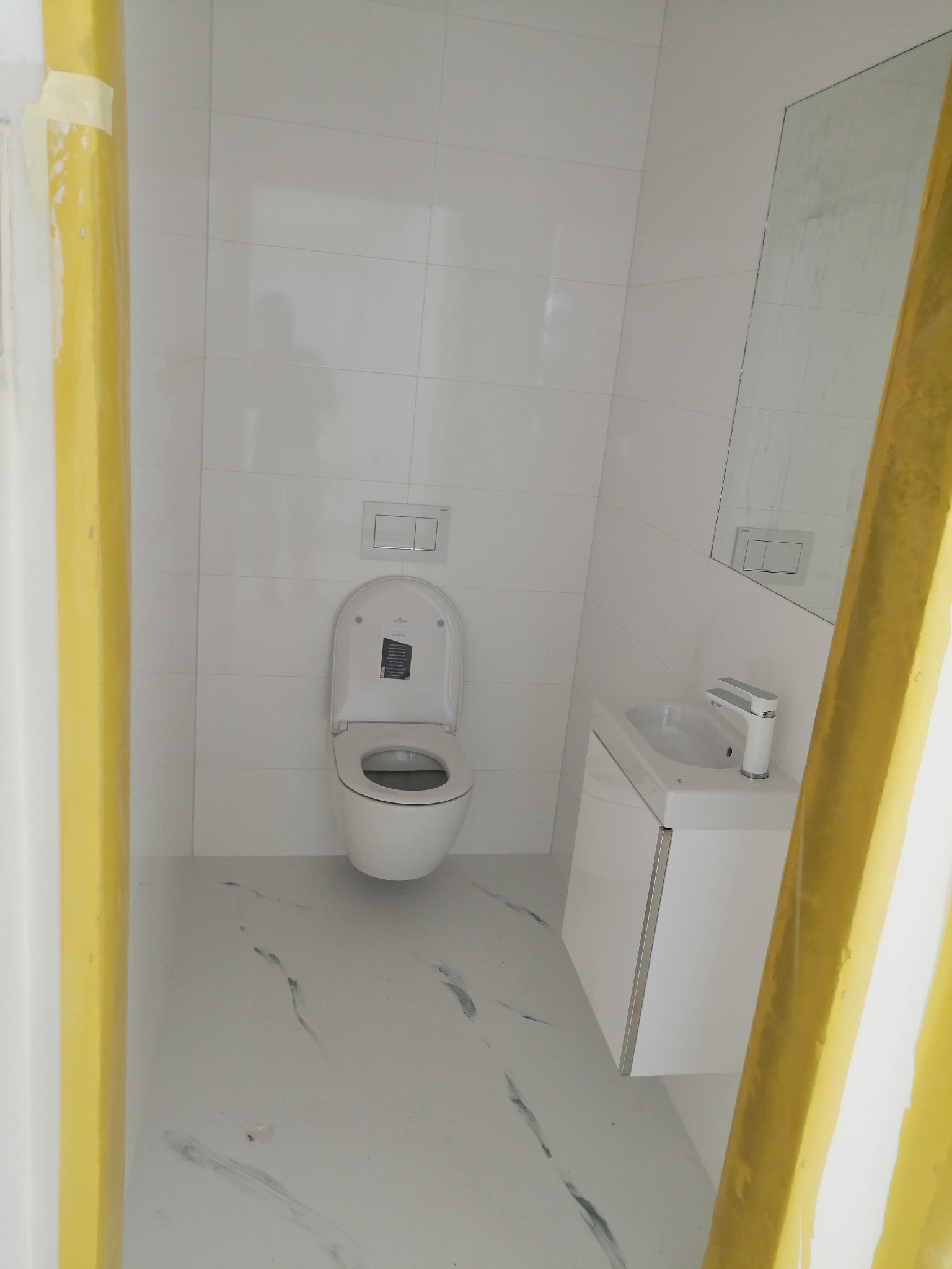 Jasna łazienka z białymi płytkami, toaleta z otwartą klapą, umywalka z szafką i lustro, widoczne elementy zabezpieczające podczas remontu.