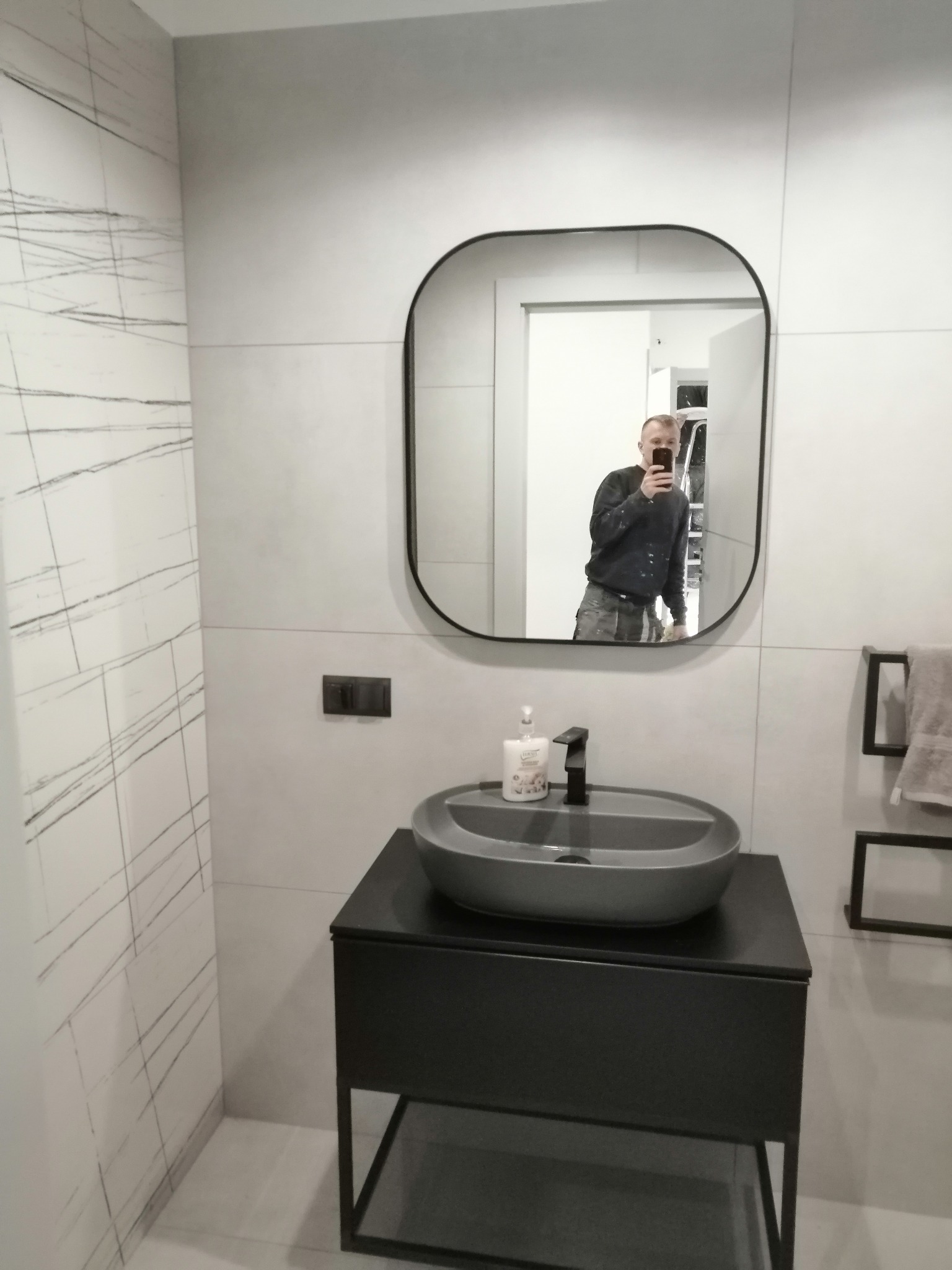 Nowoczesna łazienka z szarą umywalką nablatową i czarną armaturą, widoczna w lustrze osoba robiąca zdjęcie, ściany wyłożone płytkami imitującymi marmur.