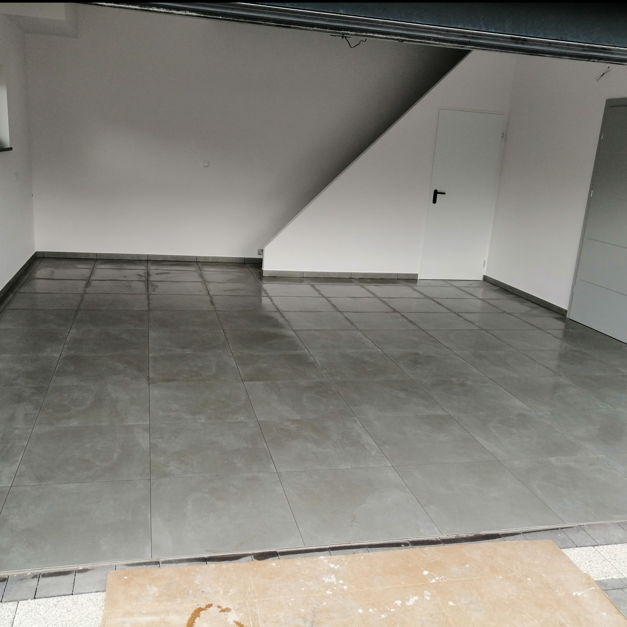 Wykończona podłoga z szarych, kwadratowych płytek w garażu lub pomieszczeniu gospodarczym. Widoczne białe ściany, białe drzwi i fragment bramy garażowej.