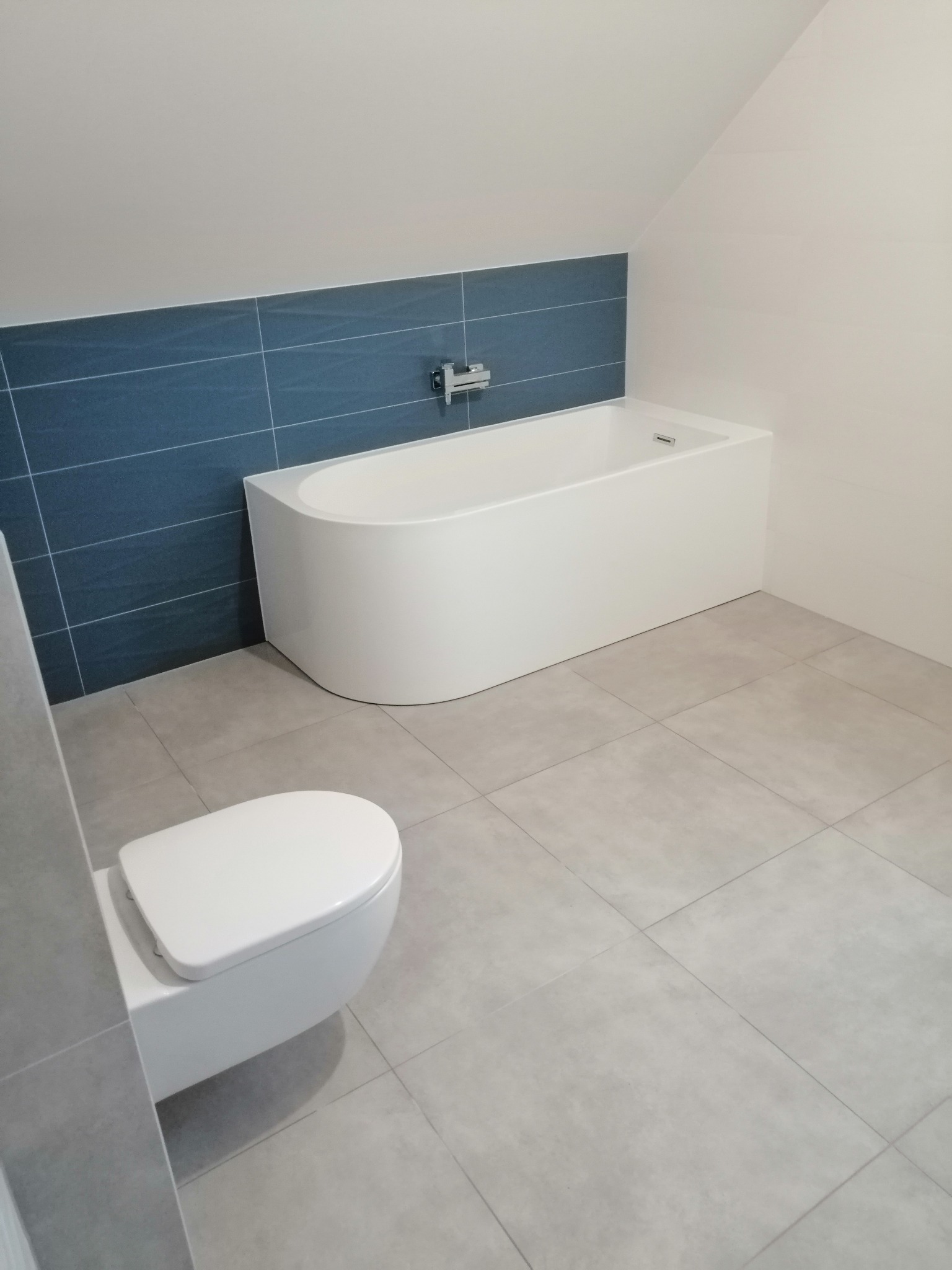 Nowoczesna, minimalistyczna łazienka na poddaszu z białą wanną zabudowaną, wiszącą toaletą i szarymi płytkami podłogowymi oraz pasem ciemnoniebieskich płytek na ścianie.