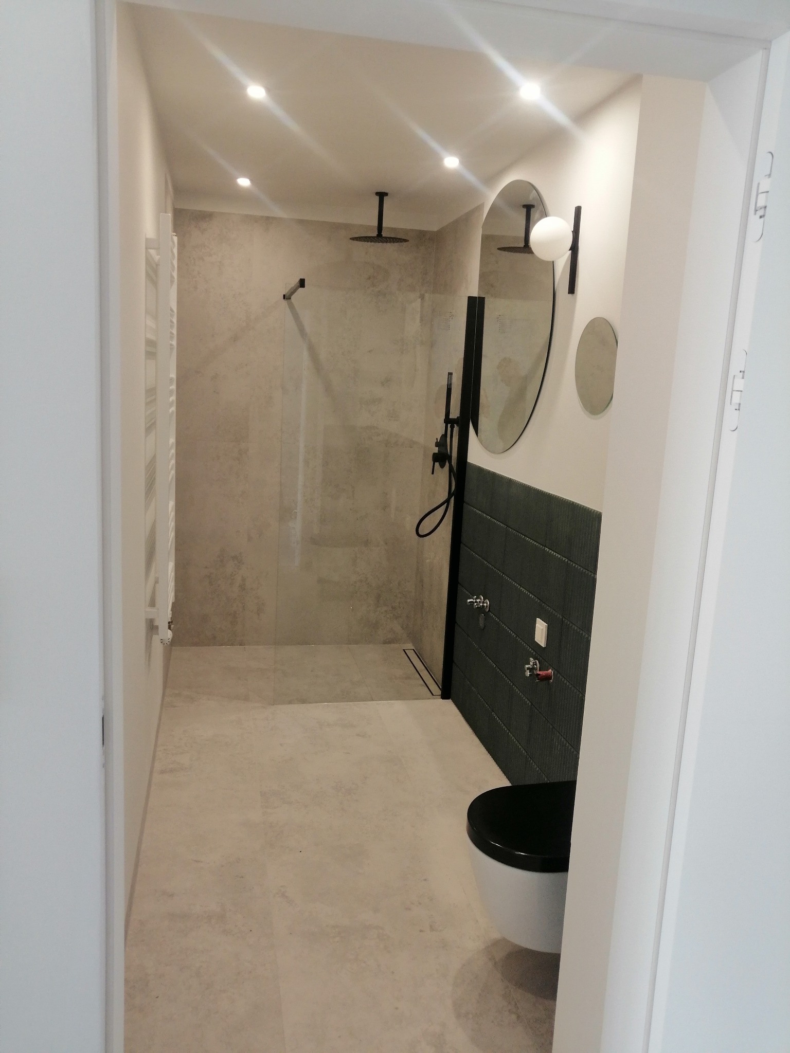 Nowoczesna łazienka z kabiną prysznicową typu walk-in, szarą glazurą imitującą beton, czarną armaturą i wiszącą toaletą z czarną deską, oświetlona punktowymi lampami sufitowymi.