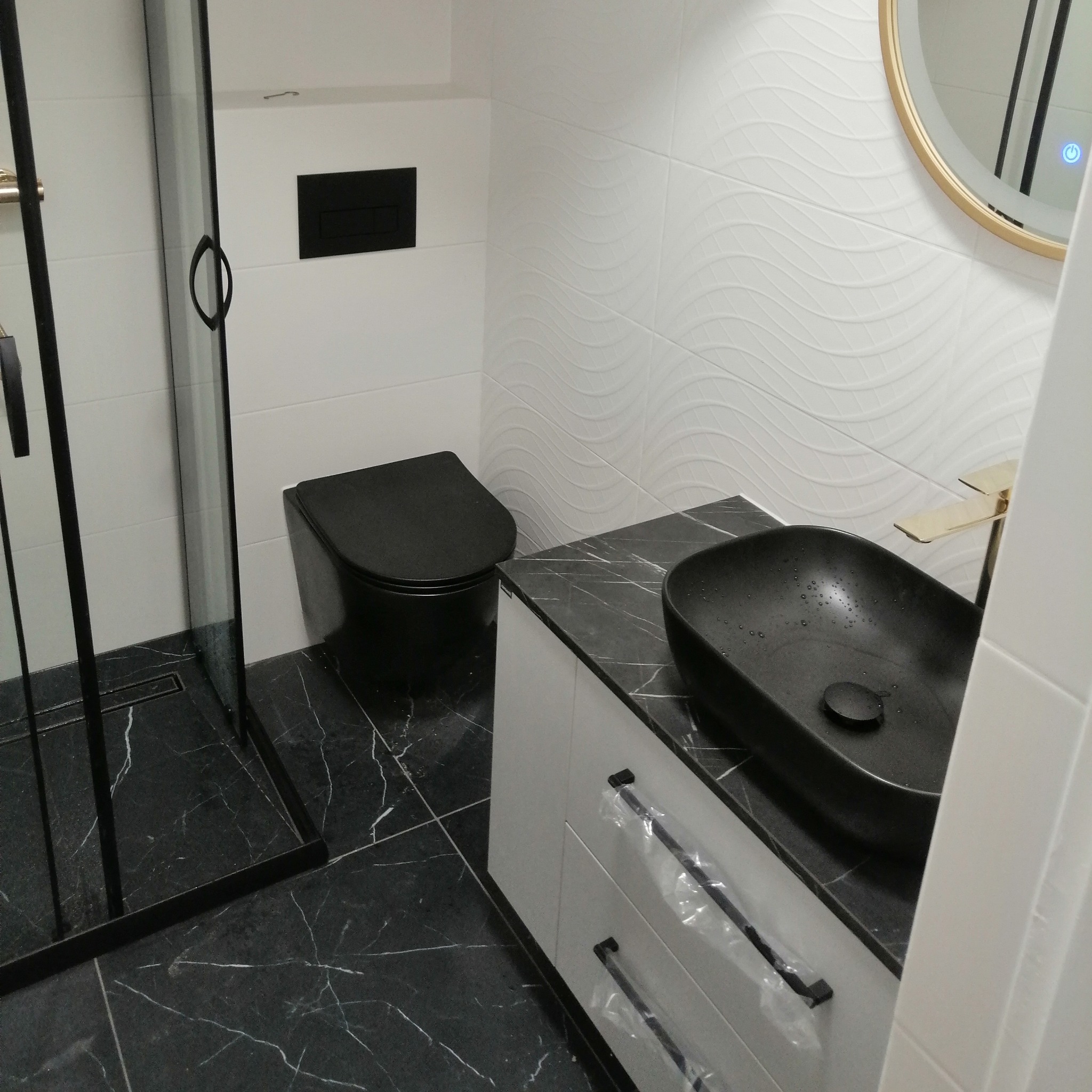 Nowoczesna łazienka z czarną ceramiką: toaleta, umywalka na blacie imitującym marmur, złota bateria i kabina prysznicowa z czarnymi ramami.