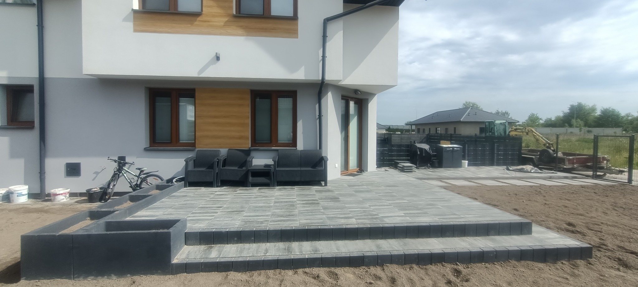 Nowoczesny taras z szarej kostki brukowej, schody z obrzeżami, meble ogrodowe na tle domu, w tle częściowo widoczny sprzęt budowlany.
