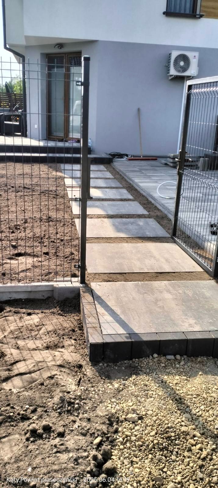 Nowoczesna ścieżka z płyt betonowych w ogrodzie, obramowana czarną kostką i ogrodzeniem panelowym. Widok na budynek z klimatyzatorem i otwartą bramką.