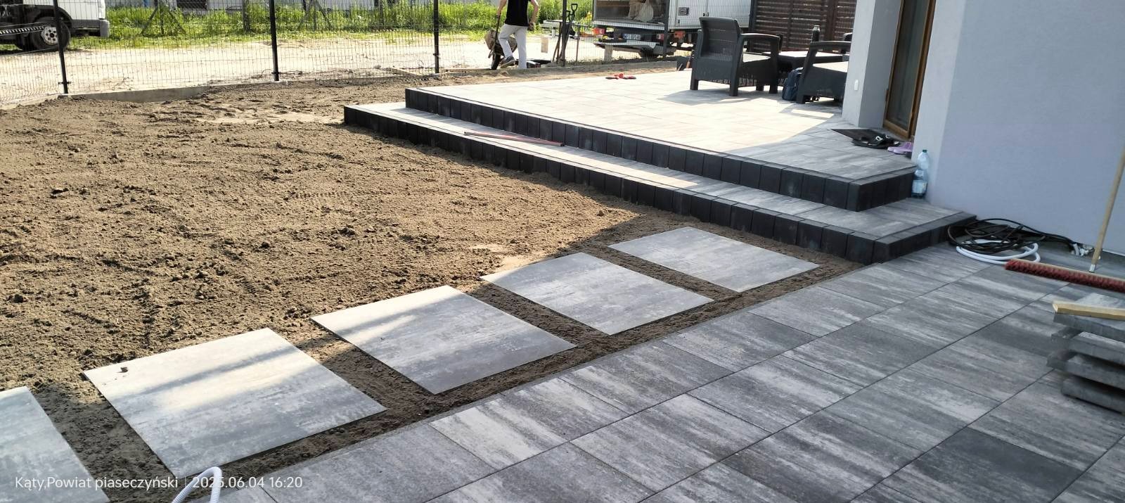 Etap układania szarej kostki brukowej na posesji. Część kostki ułożona, część w trakcie układania. Widoczne przygotowane podłoże i narzędzia.