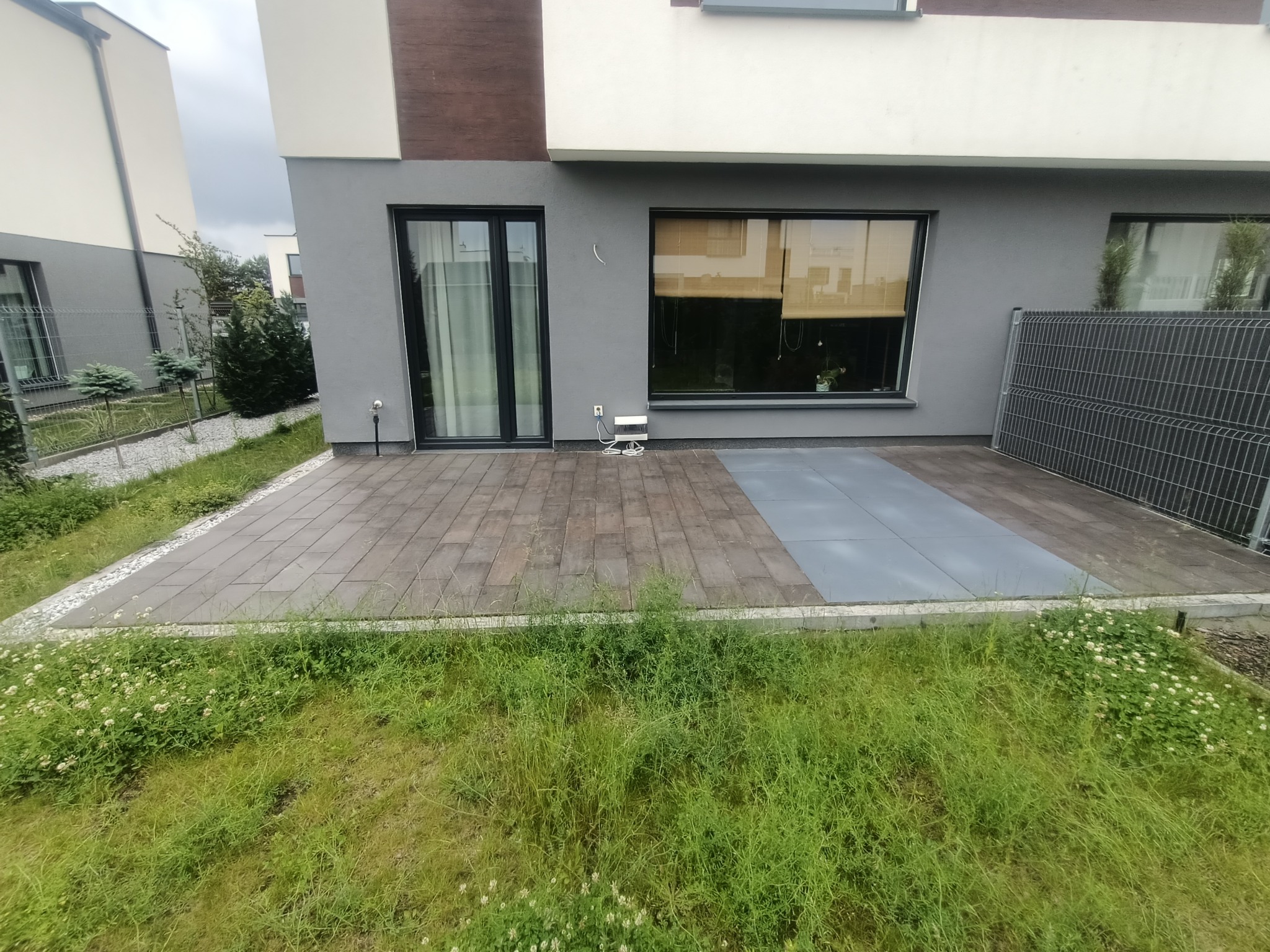 Nowoczesny taras z kostki brukowej w dwóch odcieniach szarości przylegający do domu z dużymi oknami i fragmentem panelowego ogrodzenia.