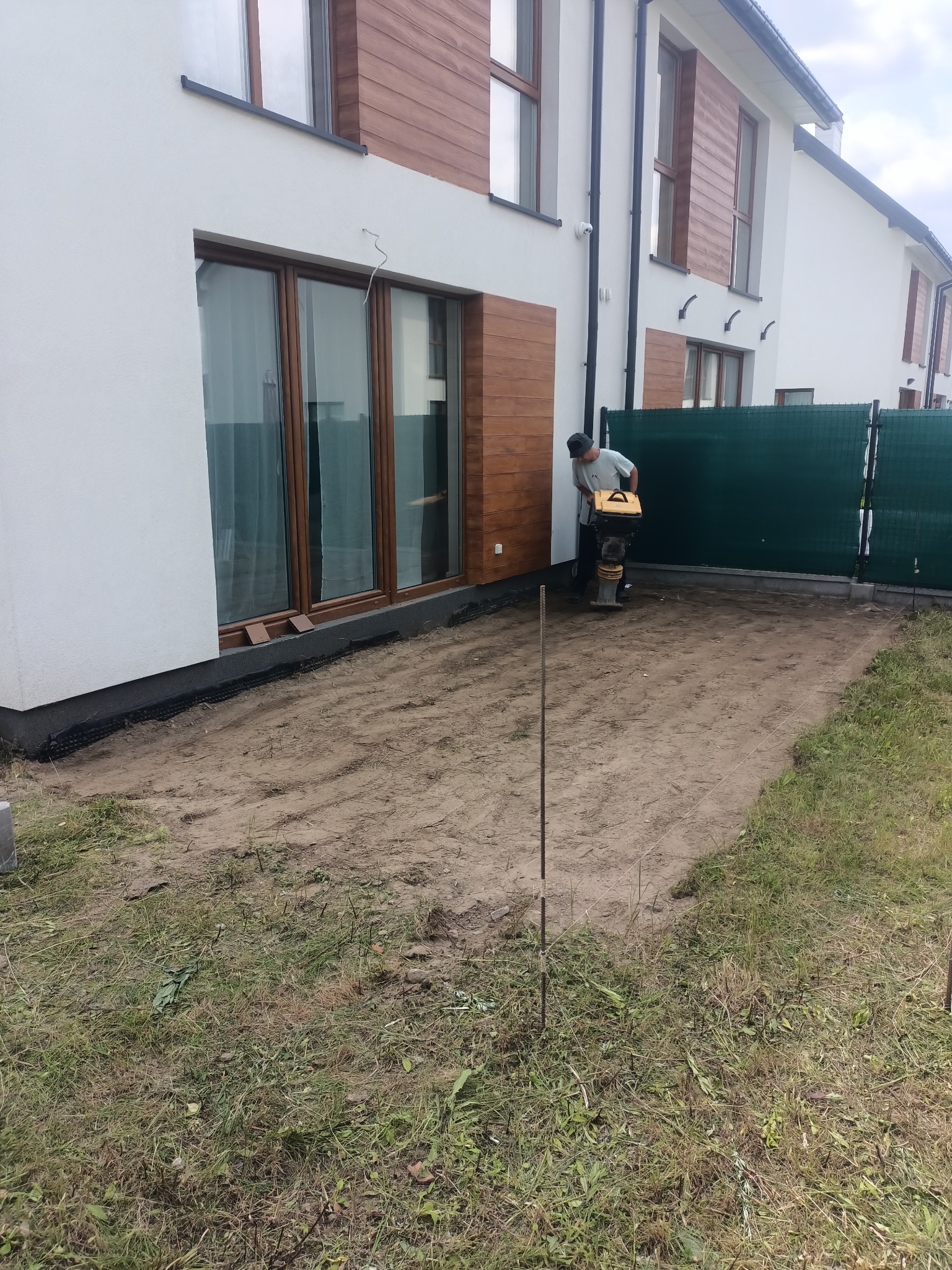 Prace przygotowawcze pod kostkę brukową: ubijanie ziemi zagęszczarką przy nowoczesnym domu z drewnianą elewacją. Widoczne obrzeża i wstępnie wyrównany teren.