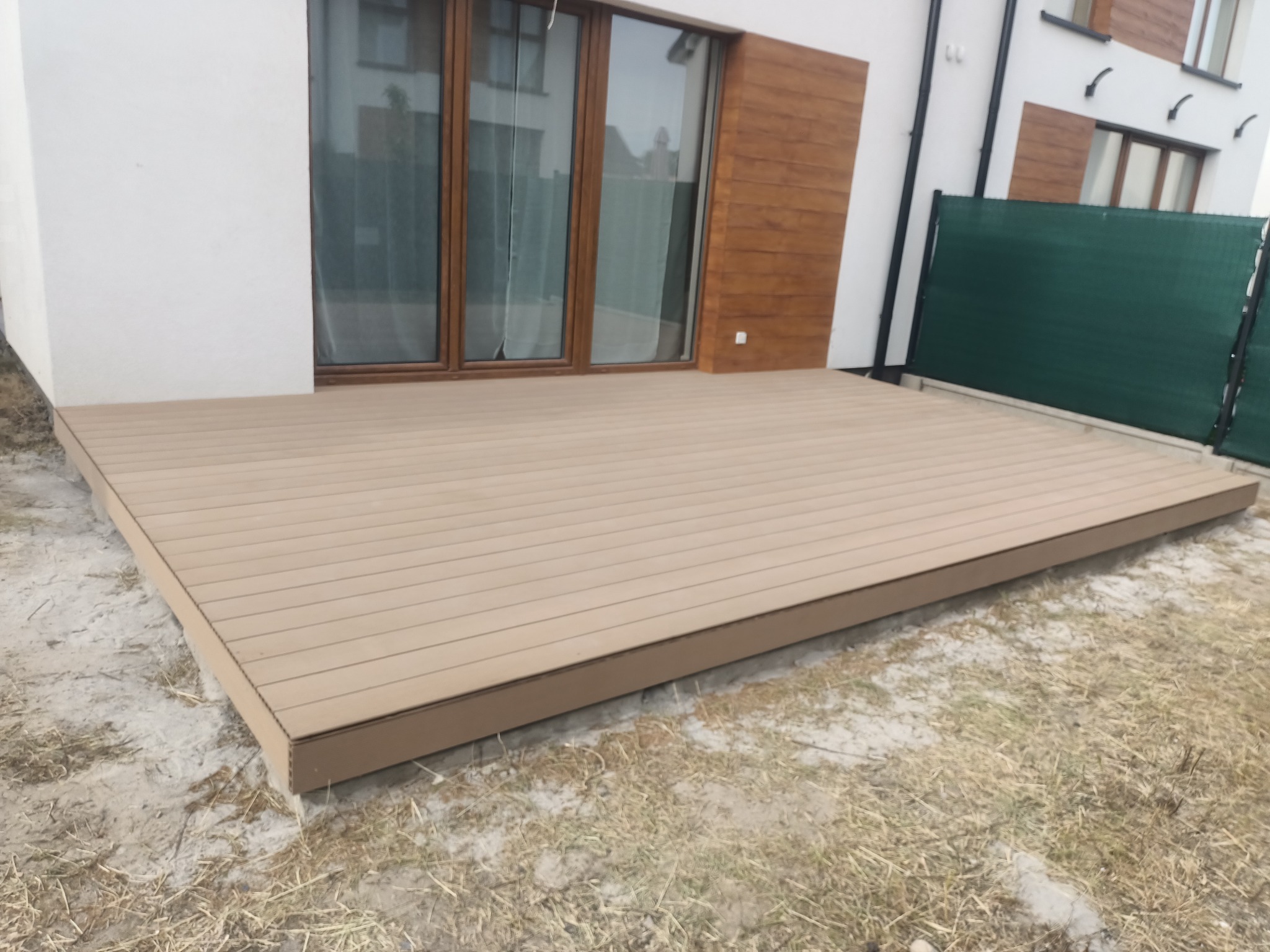 Nowoczesny taras z deski kompozytowej w kolorze jasnego drewna, przylegający do białego budynku z drewnianymi akcentami. Widok na zielone ogrodzenie.