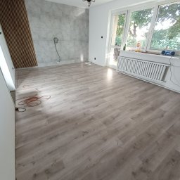 Bigbud - Wnętrze pokoju po remoncie z panelami ściennymi imitującymi beton i dekoracyjnymi listwami z drewna, widoczne kable wystające ze ściany, parkiet na podłodze i grzejnik pod oknem.