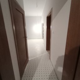 Bigbud - Wykończony korytarz z jasnymi ścianami, wzorzystą podłogą i drewnianymi drzwiami, widok z perspektywy wnętrza, kabel zwisający z sufitu.