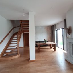 Bigbud - Jasne, przestronne wnętrze domu z drewnianymi schodami, parkietem i stołem. Widoczne niedokończone punkty oświetleniowe na suficie.