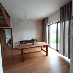 Bigbud - Jasne wnętrze domu z drewnianą podłogą, stołem z bukietem tulipanów, ścianą z motywem roślinnym i szarymi zasłonami przy dużym oknie.