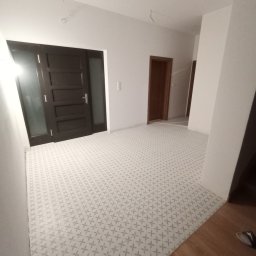 Bigbud - Wnętrze nowo wykończonego przedpokoju z białą ścianą, ciemnymi drzwiami wejściowymi i wzorzystą podłogą w odcieniach szarości, widoczne niedokończone przewody elektryczne na suficie.