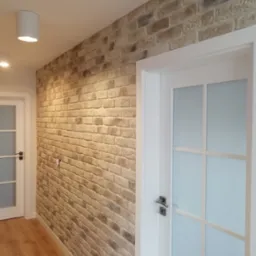 Ściana wewnętrzna wykończona dekoracyjną cegłą w odcieniach beżu i szarości, sąsiadująca z białą, przeszkloną futryną drzwiową, oświetlona nowoczesnym plafonem.