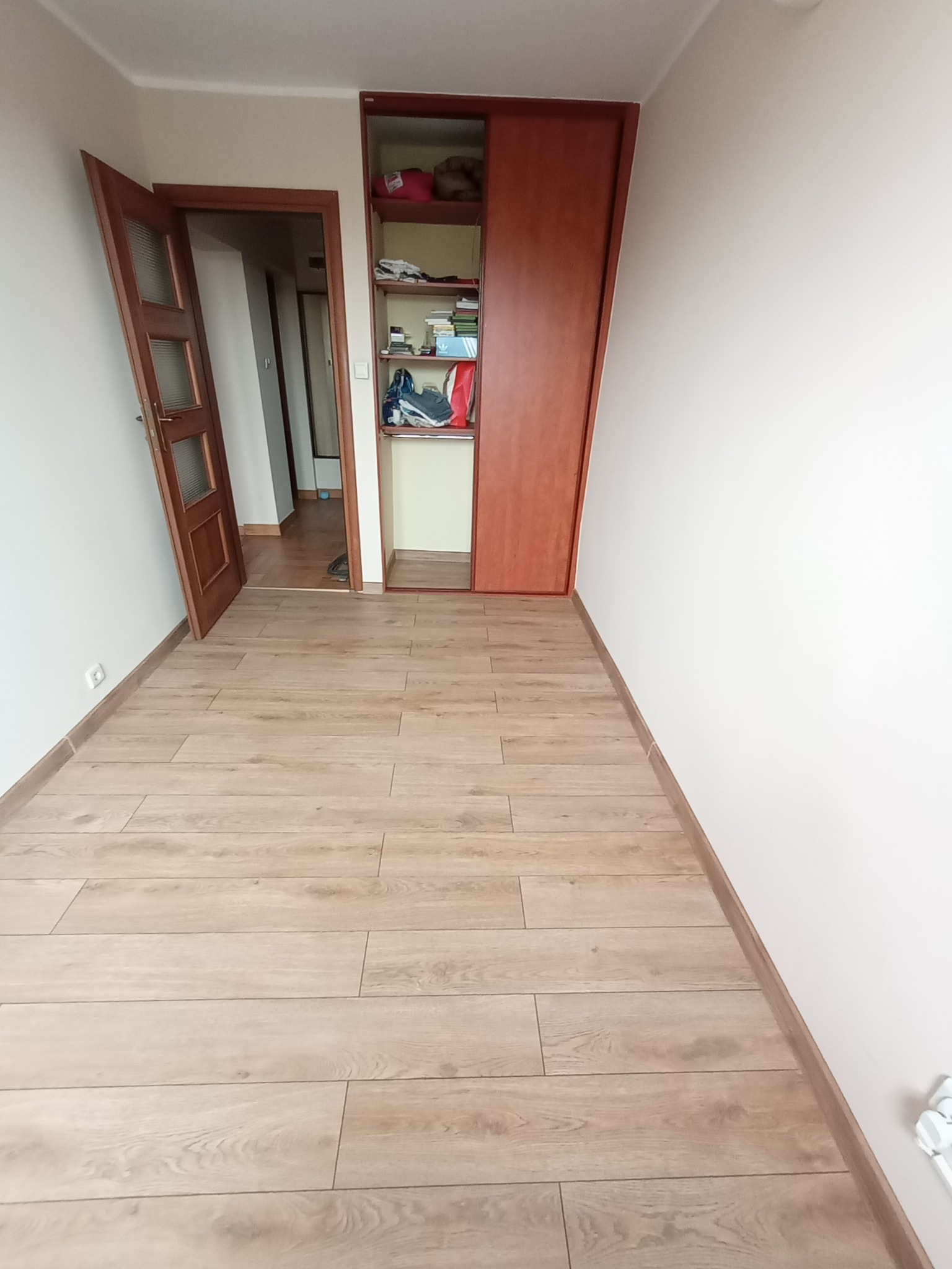 Pusty pokój po remoncie z drewnianą podłogą, otwartymi drzwiami i szafą wnękową z częściowo otwartymi drzwiami przesuwnymi.