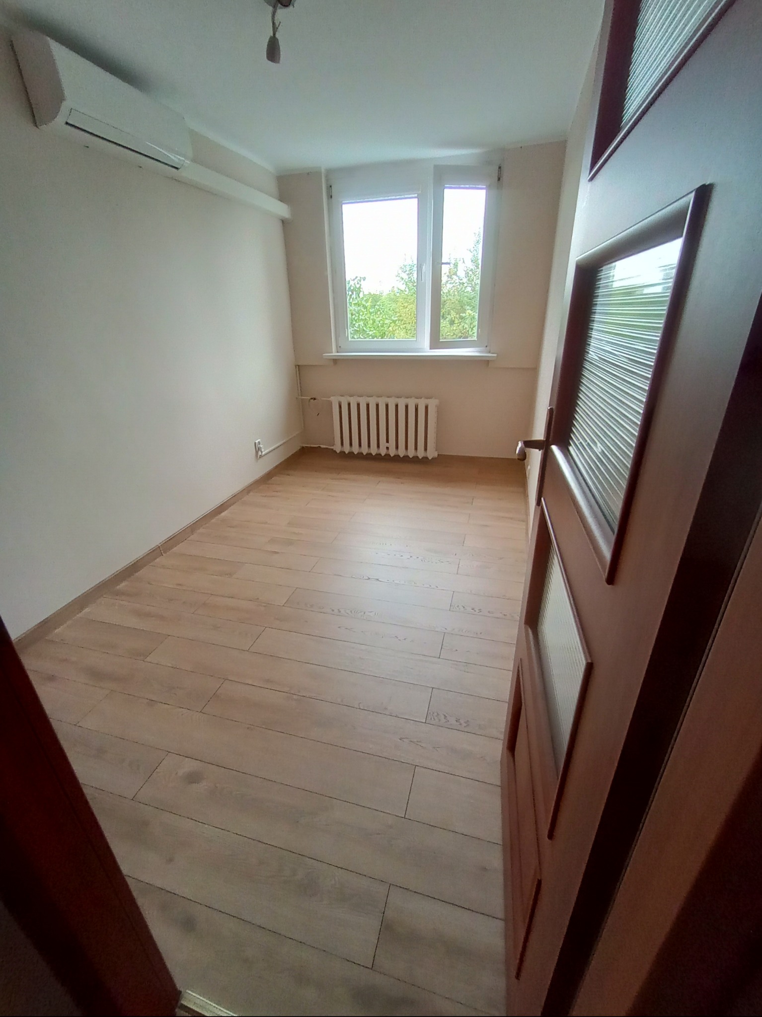 Pusty pokój z drewnianą podłogą, białymi ścianami, grzejnikiem pod oknem i klimatyzatorem na ścianie, widziany z perspektywy otwartych drzwi.